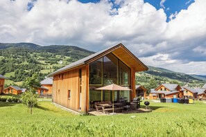 Una cabaña de madera con ventanas amplias, un patio con mobiliario exterior y vistas a las colinas verdes circundantes.