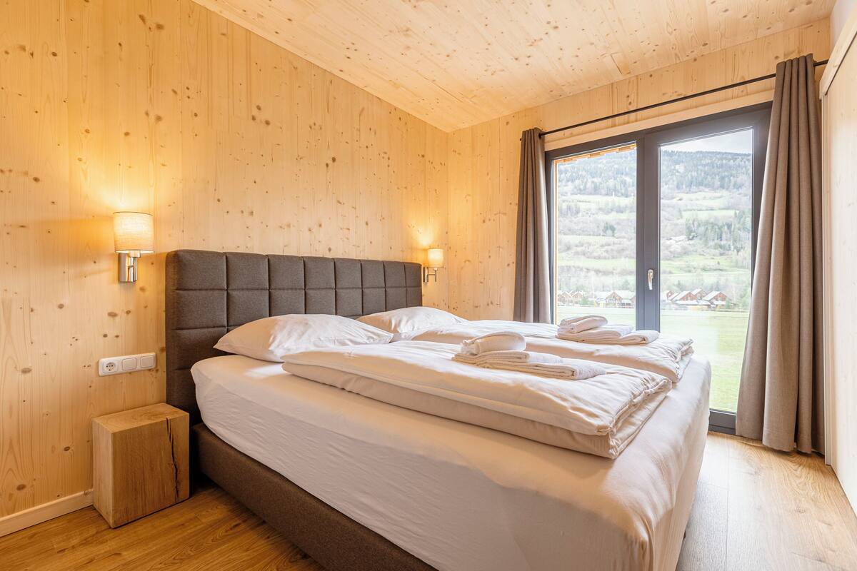 A bedroom with a bed, two bedside lamps, a wooden nightstand, and a view of a landscape through a sliding door.