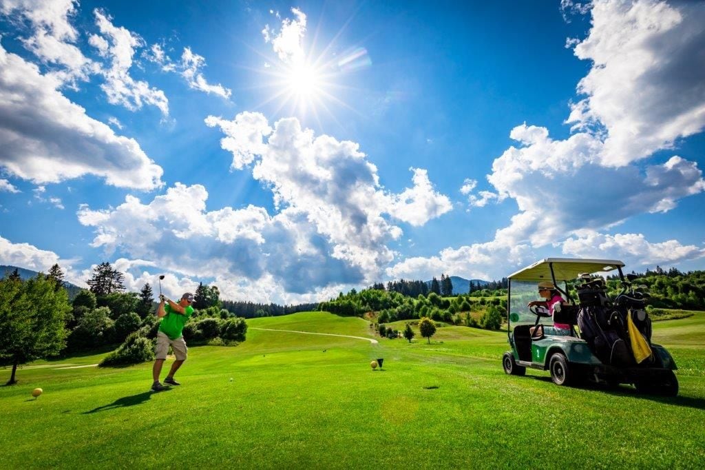 Ein Golfplatz bei blauem Himmel, ein Golfer im Schwung und ein Golfwagen.