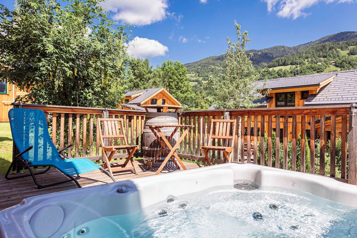 Una terraza de madera con un jacuzzi, asientos exteriores y vistas a un paisaje montañoso.