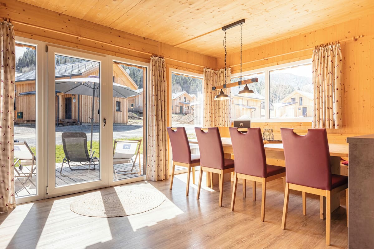 Interior de madera con comedor, ventanas amplias y vistas a cabañas exteriores.
