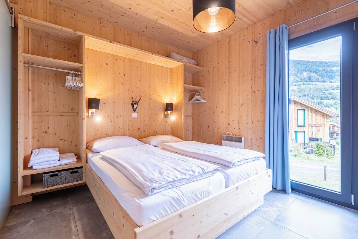 Habitación de madera con una cama, dos luces fijadas a la pared, un estante con toallas y vistas a edificios a través de un ventanal grande.