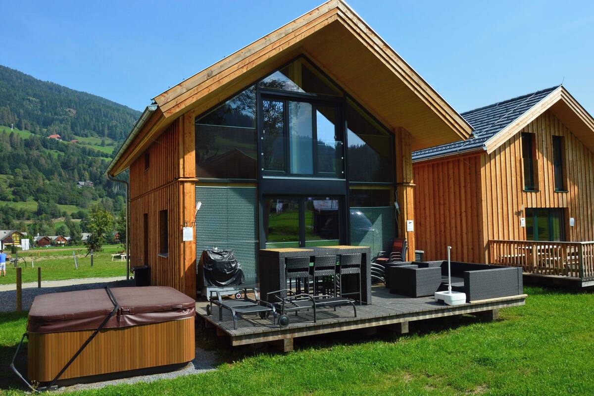 A wooden cabin with large windows, a deck with outdoor furniture, and a hot tub.