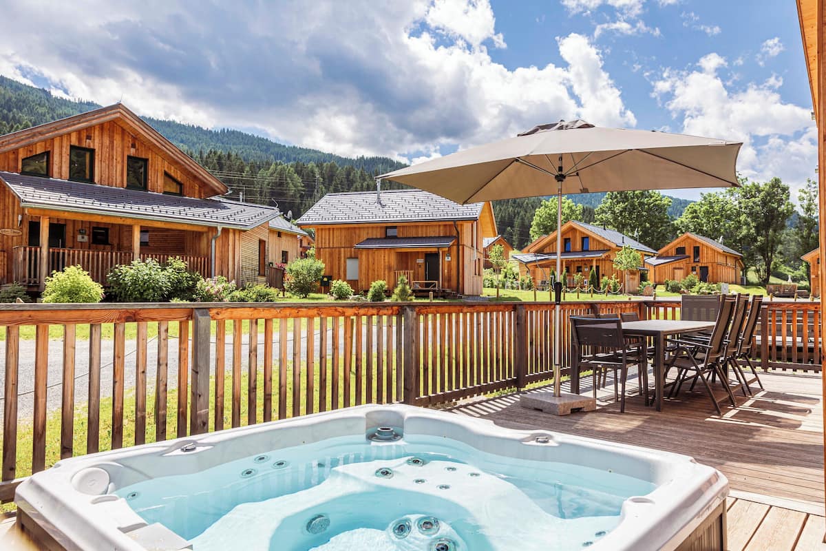 Una cabaña de madera con un jacuzzi en una terraza, rodeada por una valla de madera y un paisaje montañoso.