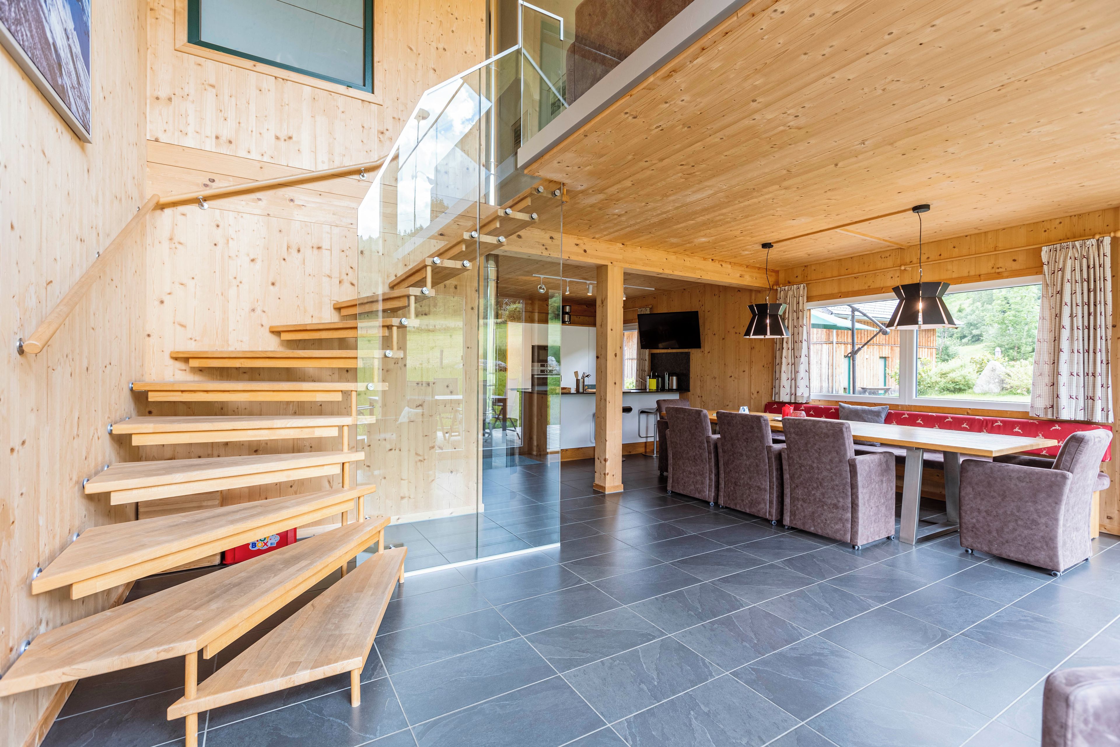 A modern interior with a wooden staircase, a dining area with chairs and a table, and a kitchen in the background.