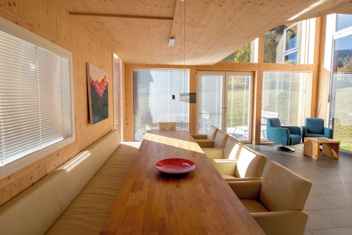 Un interior moderno con techo de madera, ventanas amplias y una mesa de madera central con un cuenco rojo.