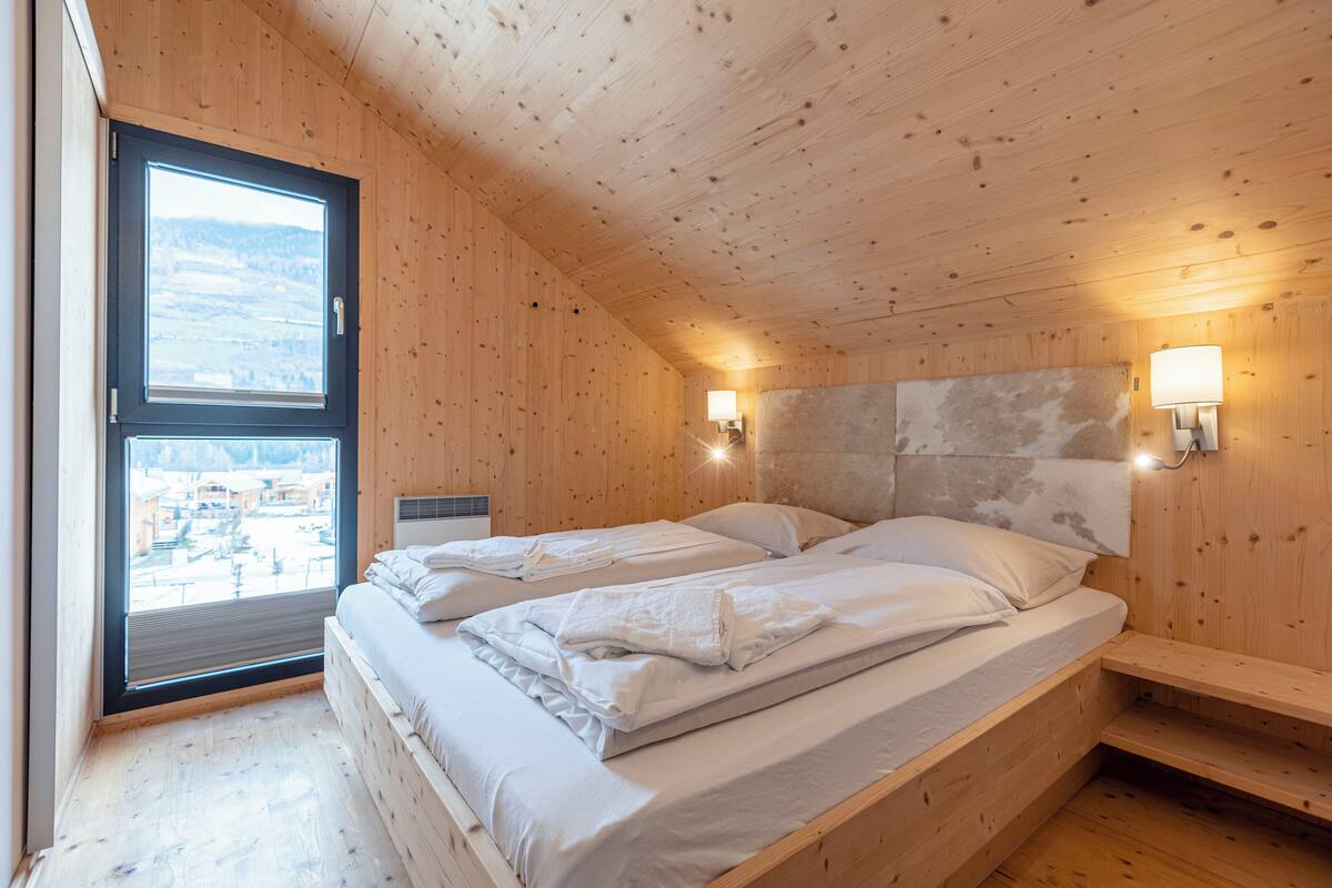 Un dormitorio de cabaña de madera con dos camas, un ventanal grande y dos lámparas fijadas a la pared.