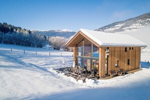 Una cabaña de madera con techo nevado, ventanas grandes y un comedor con sillas y mesas.