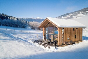 Una cabaña de madera con techo nevado, ventanas grandes y un comedor con sillas y mesas.