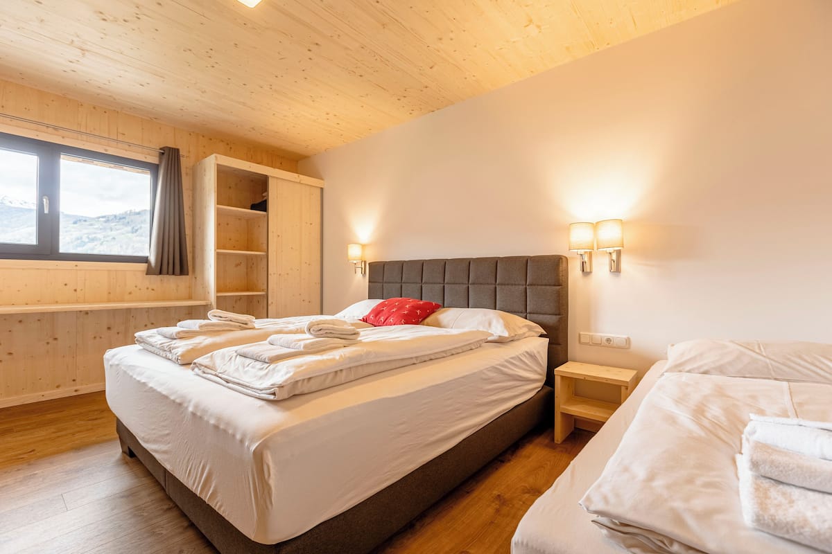A bedroom with a large bed, a bedside table, a nightstand, a window with curtains, and a wooden ceiling.