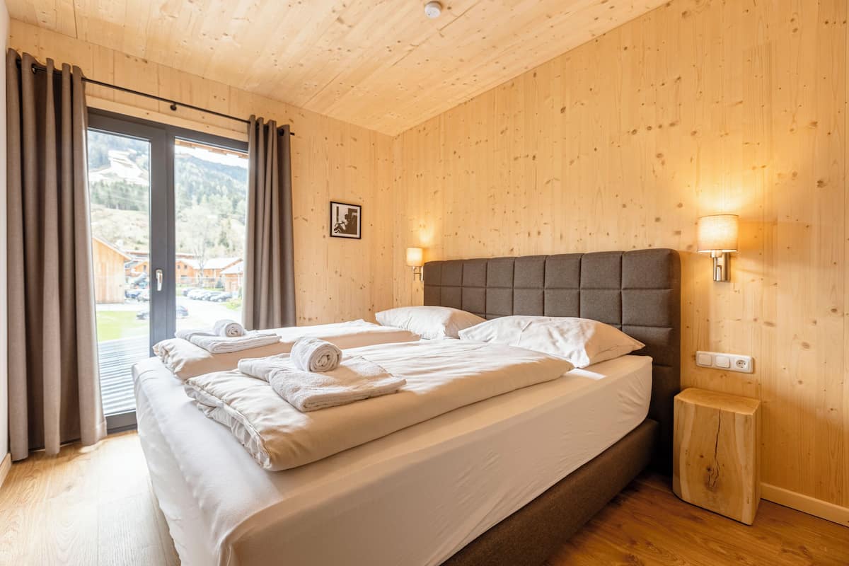 A bedroom with a wooden ceiling, a large bed with white linens, two bedside lamps, and a view of a mountain landscape through a sliding door.