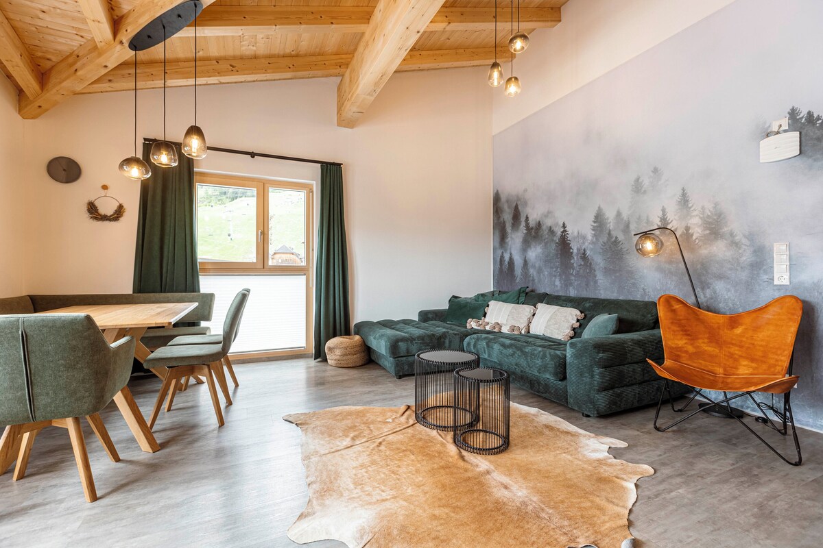 A cozy living room with a wooden ceiling, a green sofa, a dining table with chairs, and a cowhide rug.