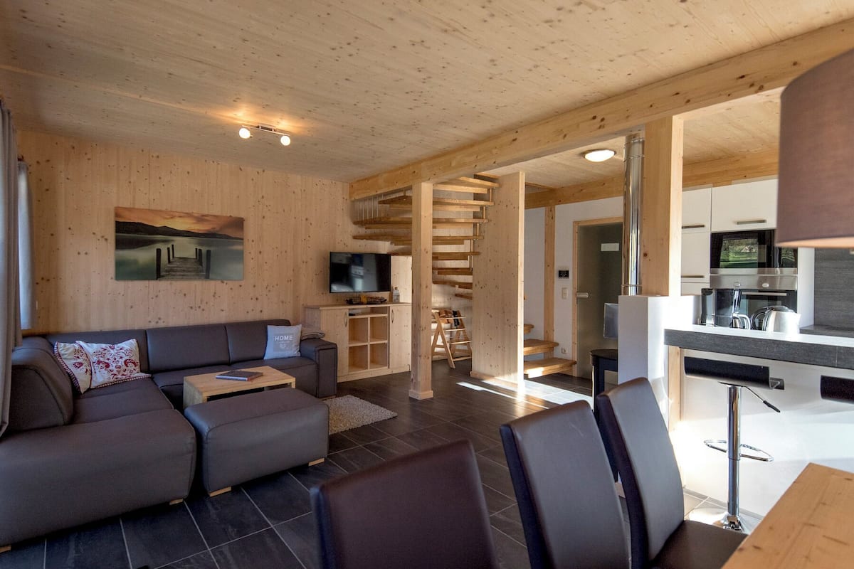 A modern living room with a sofa, a coffee table, and a TV. There is a staircase leading to a kitchen area with a dining table and chairs.