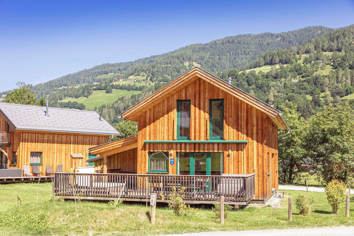 A wooden cabin with a balcony, surrounded by a grassy area and a mountain backdrop.