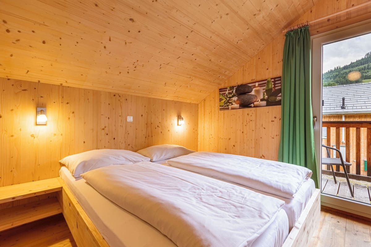 A wooden cabin room with two beds, a green curtain, and a view of a wooden deck and mountains.