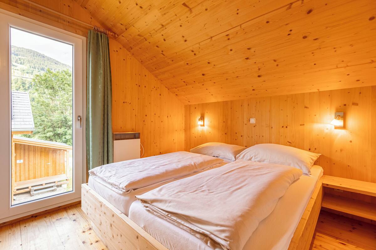 Habitación tipo cabaña de madera con dos camas, una ventana que ofrece vistas a una terraza de madera y follaje, y luces fijadas a la pared.