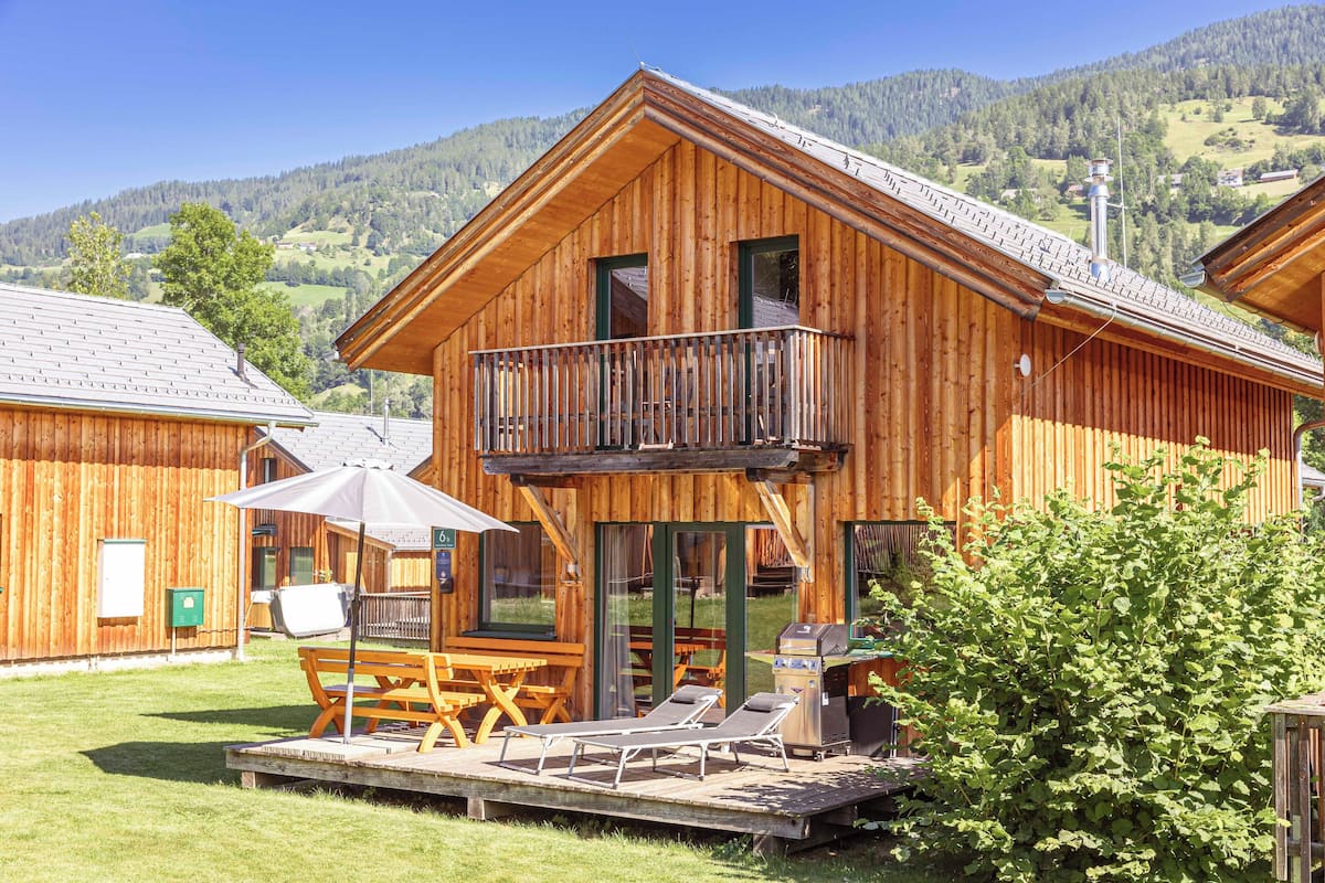 A wooden cabin with a balcony, a barbecue grill, and a poolside area with lounge chairs.