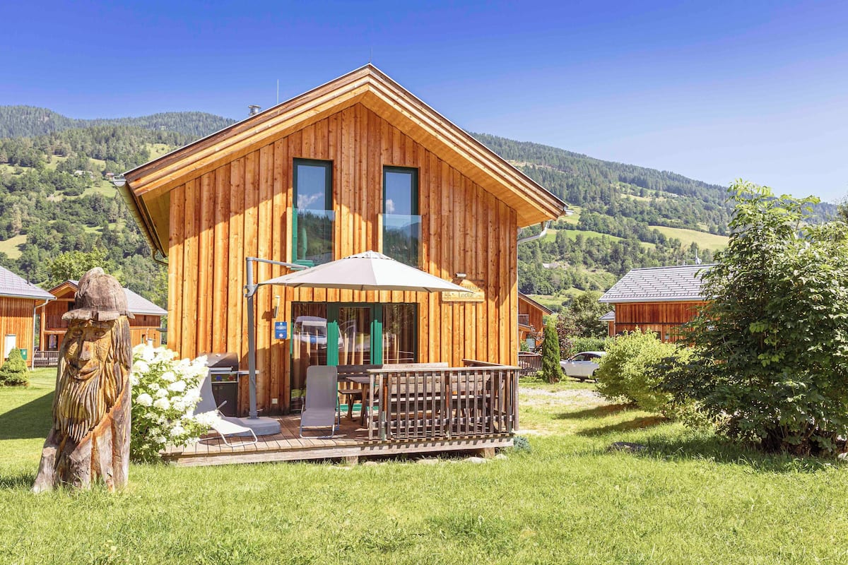 A wooden cabin with a deck, large windows, and a view of the surrounding hills.