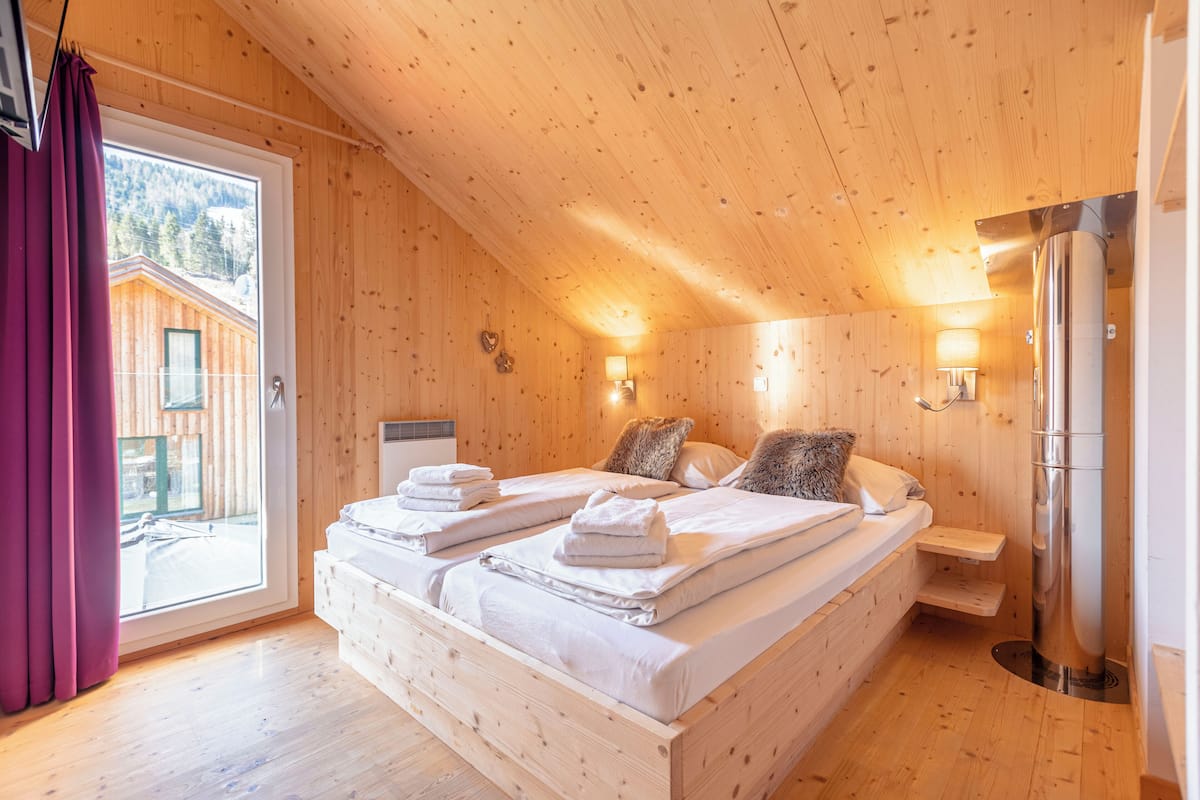 A wooden cabin bedroom with a large bed, two bedside tables, a window with curtains, and a wooden exterior visible through the window.