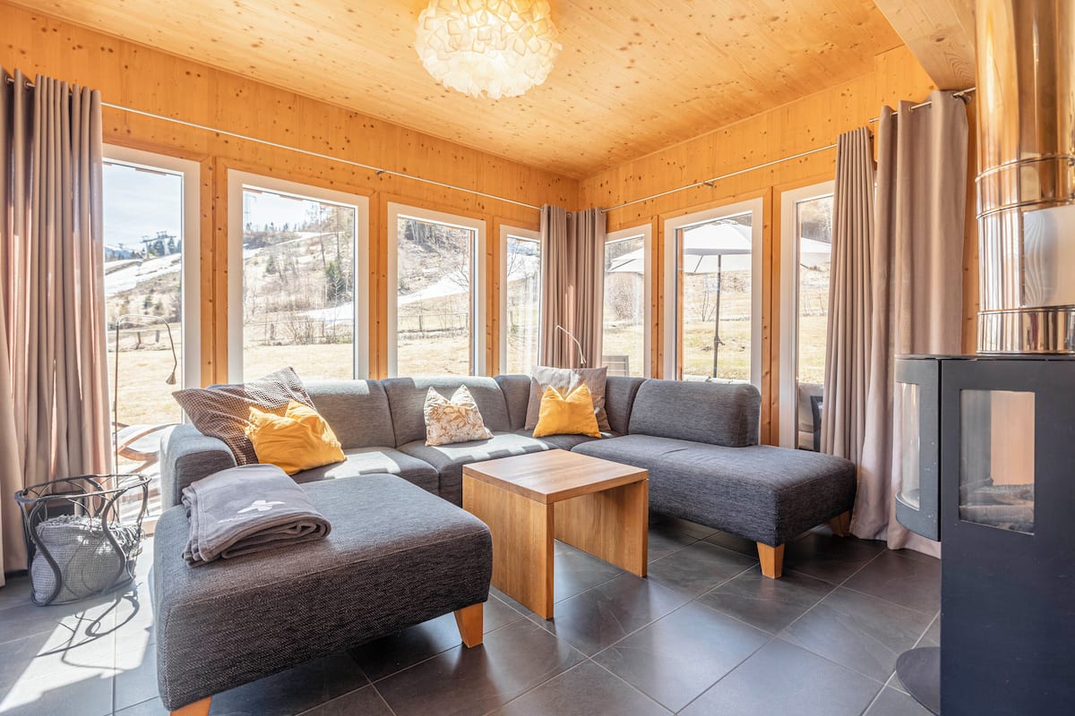 Una sala moderna con un sofá seccional, una mesita de madera y grandes ventanas que ofrecen vistas al exterior.