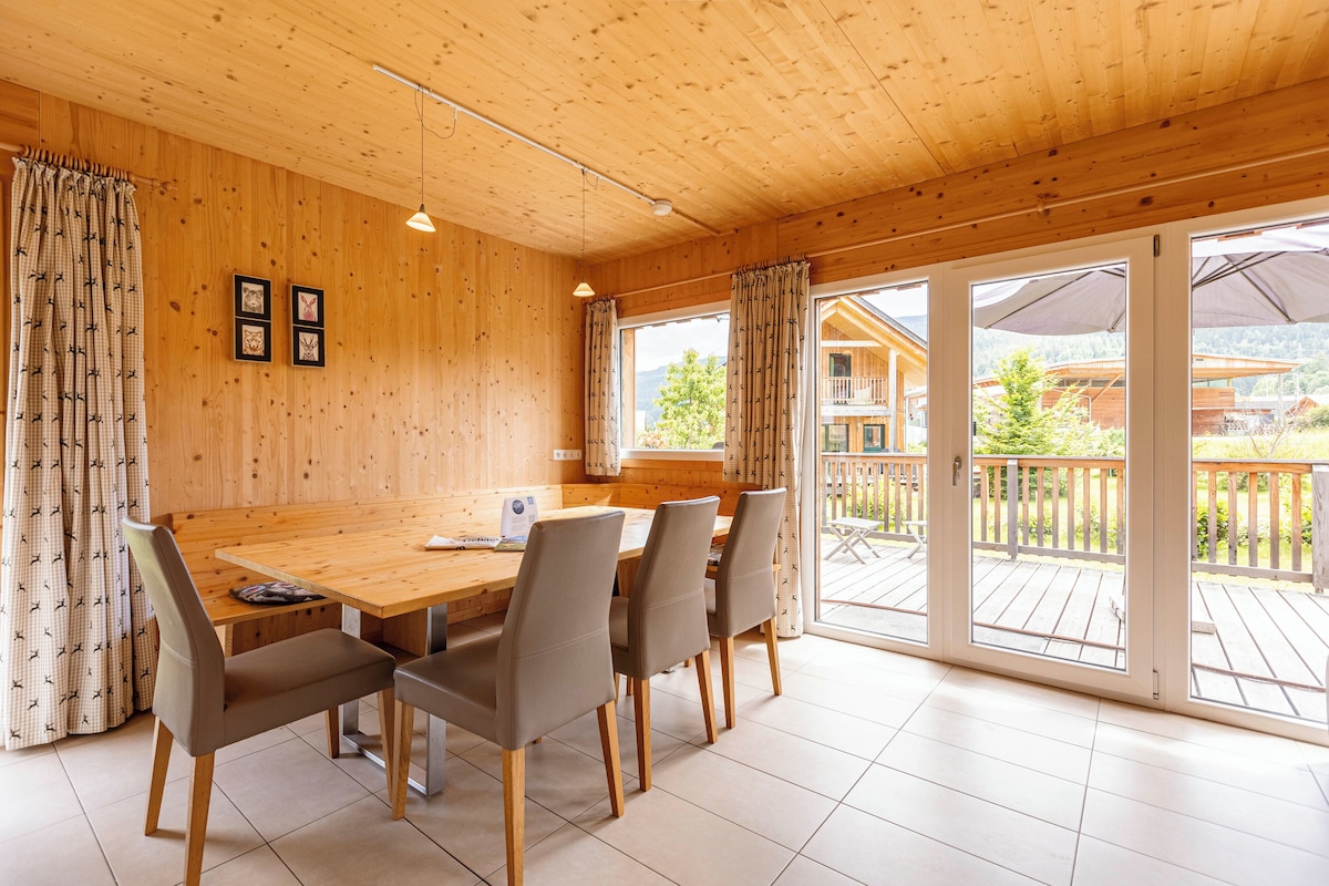 A wooden interior with a dining table and chairs, large windows, and a view of a wooden building and mountains.