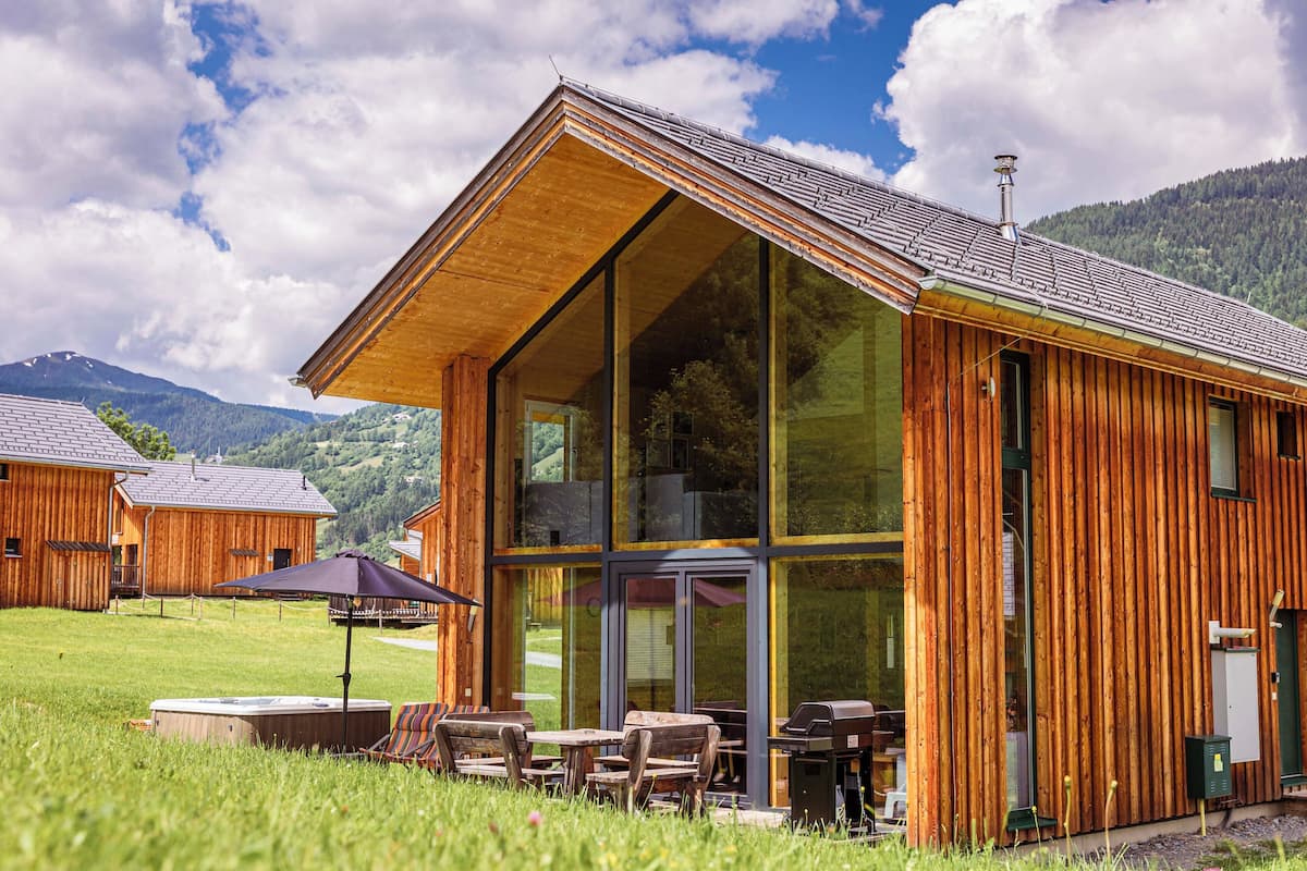 A wooden cabin with large windows, outdoor seating, and a view of the surrounding mountains.