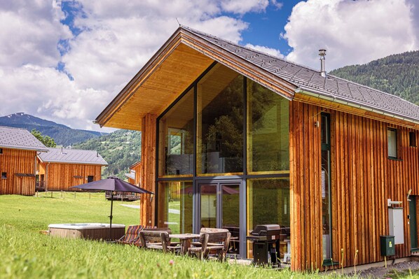 Una cabaña de madera con amplios ventanales, mobiliario exterior y vistas a las montañas circundantes.