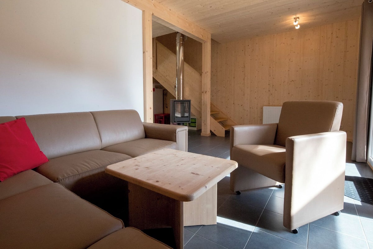 Una sala de estar moderna con un sofá, un sillón y una pequeña mesa de madera. Se ve una escalera que lleva a otro nivel y un televisor al fondo.