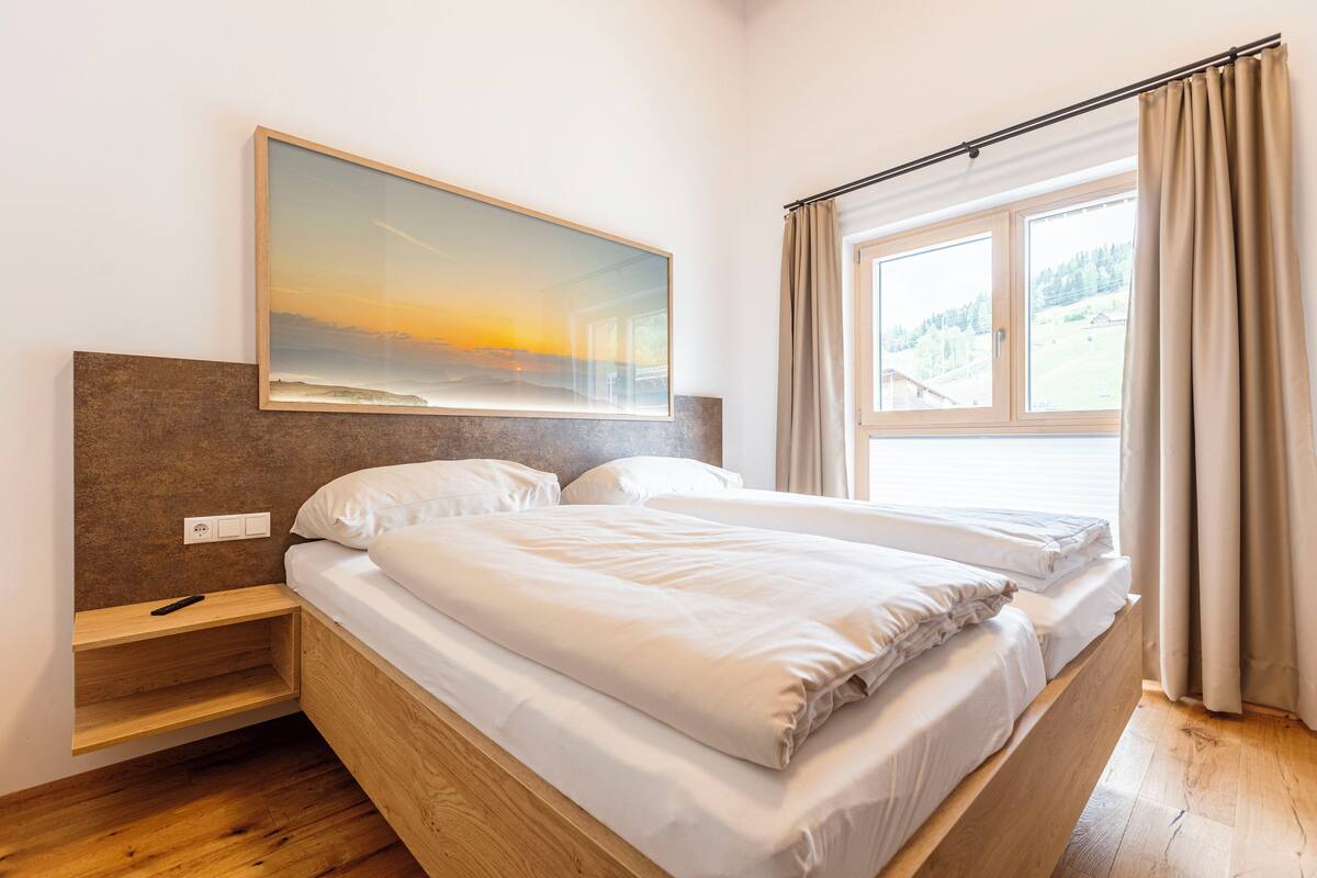A bedroom with a bed, a nightstand, a painting, and a window with curtains.
