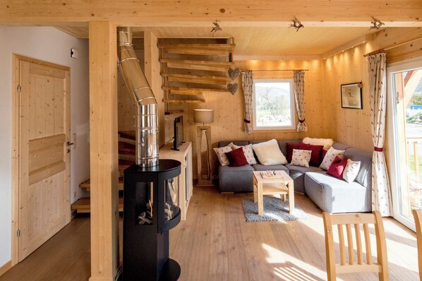 A cozy cabin interior with a central fireplace, a sofa with patterned cushions, and a wooden staircase. - Kreischberg Chalets by ALPS RESORTS (Sankt Georgen am Kreischberg)
