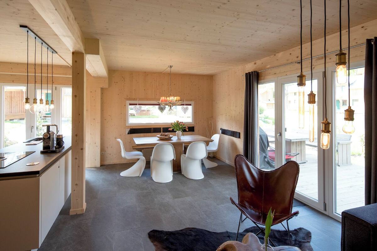 A modern kitchen with a dining area, featuring a wooden ceiling, a large window, and a mix of white and brown chairs.