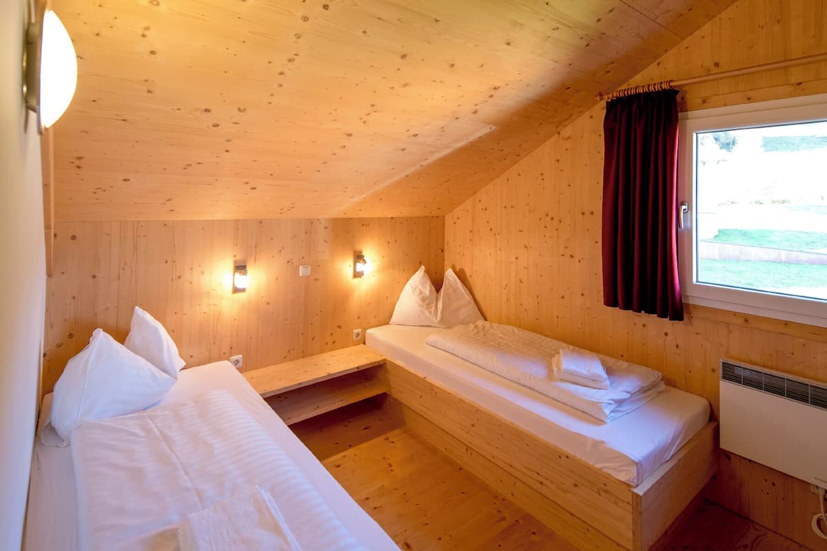 A wooden cabin room with two beds, a window with curtains, and wall-mounted lights.