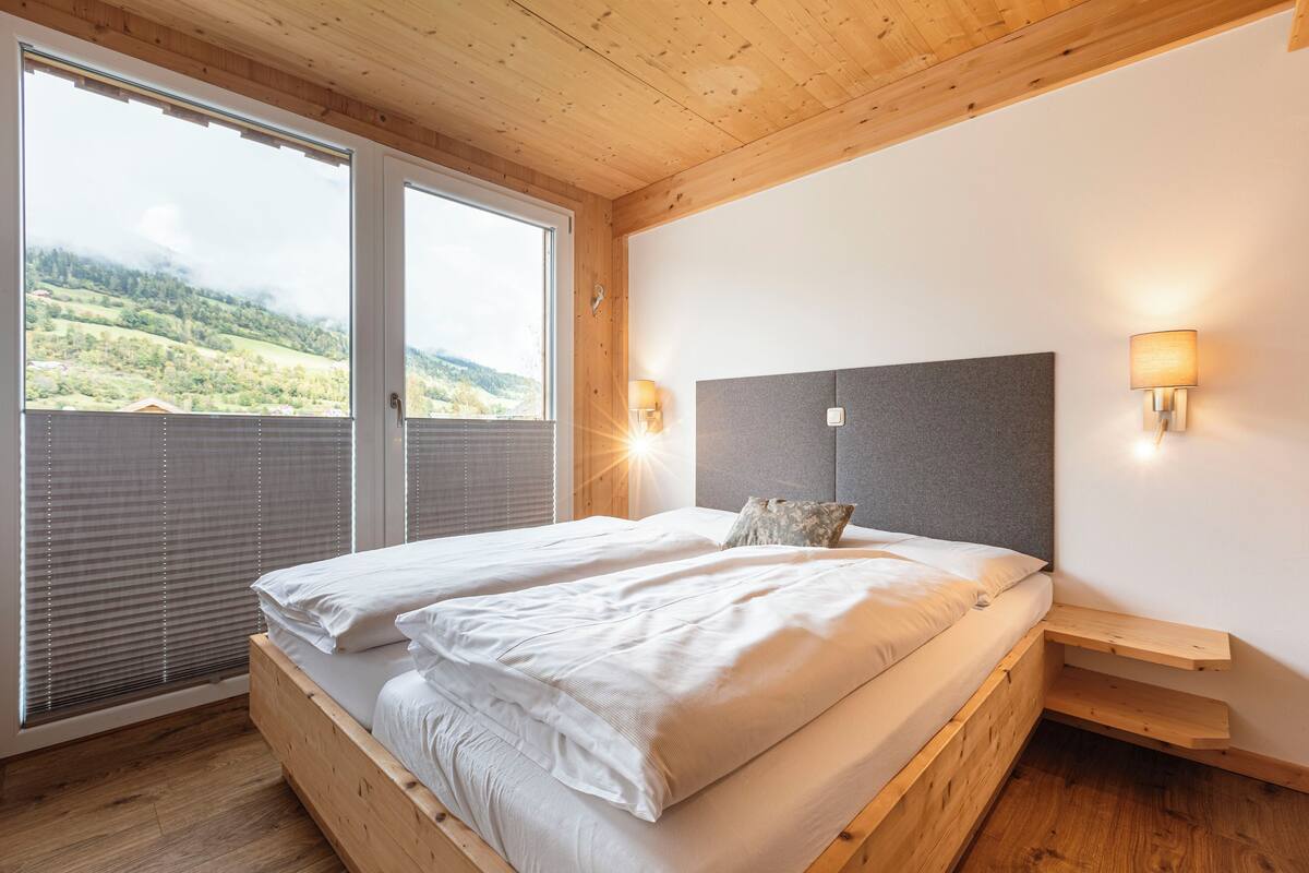 A bedroom with two beds, wooden headboard, bedside lamps, and a view of a green landscape through large windows.