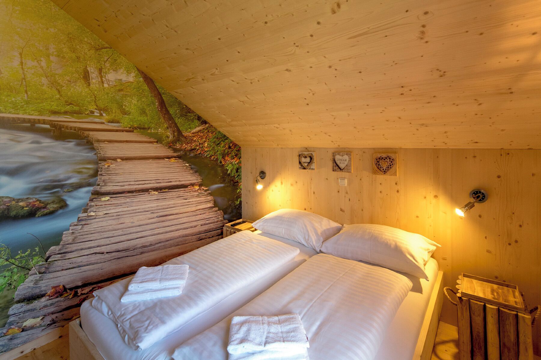 Une pièce de cabane en bois comprenant un lit, un banc en bois et donnant sur une zone boisée traversée par un ruisseau.