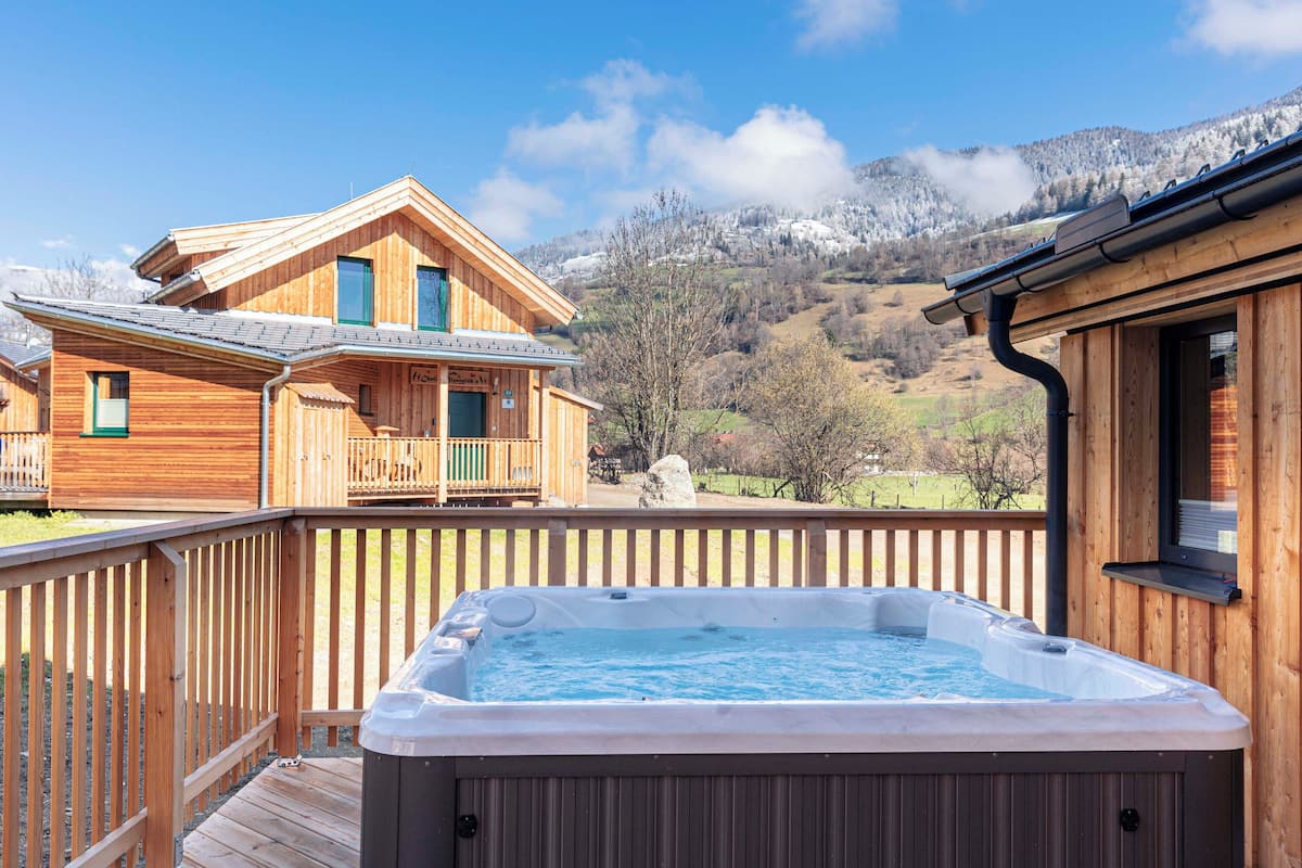 A wooden cabin with a hot tub on a deck, surrounded by mountains and a clear sky.