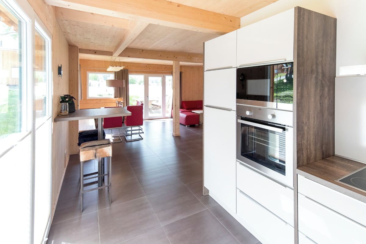Cocina moderna con armarios blancos, horno empotrado y zona de comedor con sillas rojas.