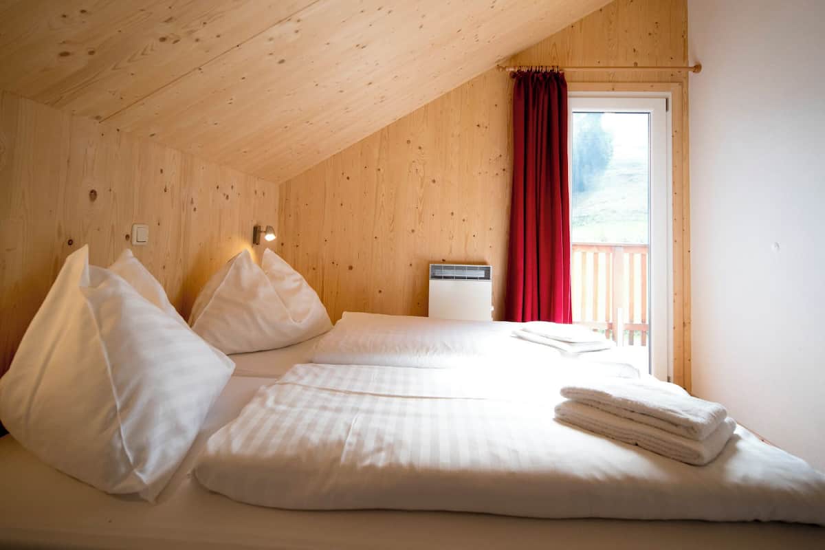 A bedroom with a wooden ceiling, a bed with white bedding, a red curtain, and a window with a view of a wooden deck.