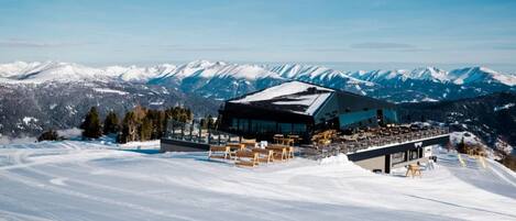 A modern ski resort building with a large terrace and snow-covered mountains in the background.
