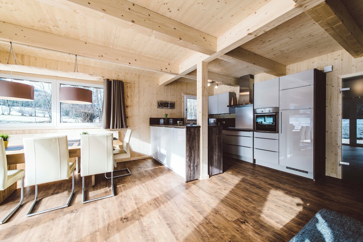 A modern kitchen with wooden cabinetry, a dining area with white chairs, and a large window providing natural light.