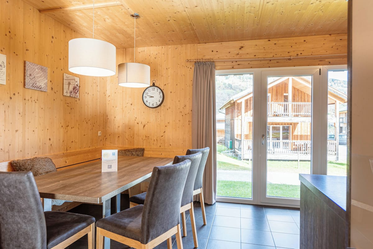 A wooden interior with a dining table, chairs, a clock, and a view of a wooden building outside.