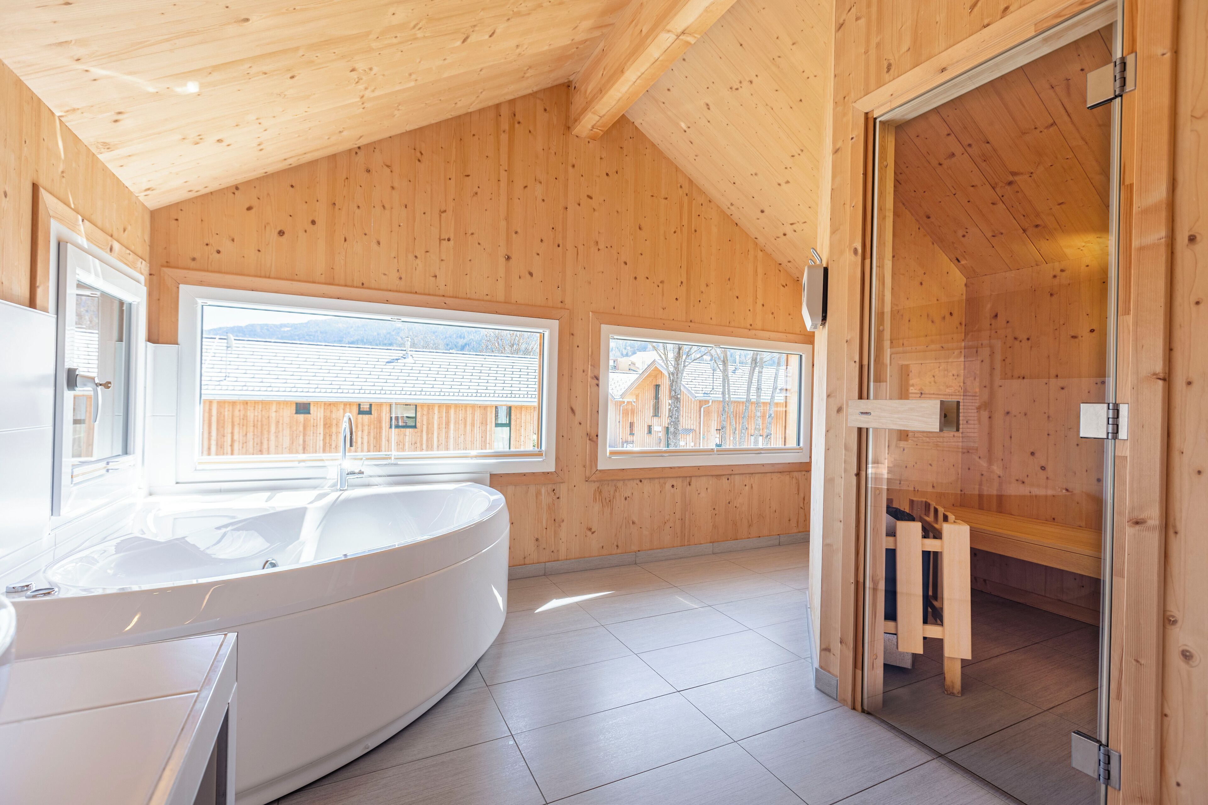 A modern wooden cabin with a large bathtub, two windows with a view of a wooden house, and a glass door leading to a shower area.