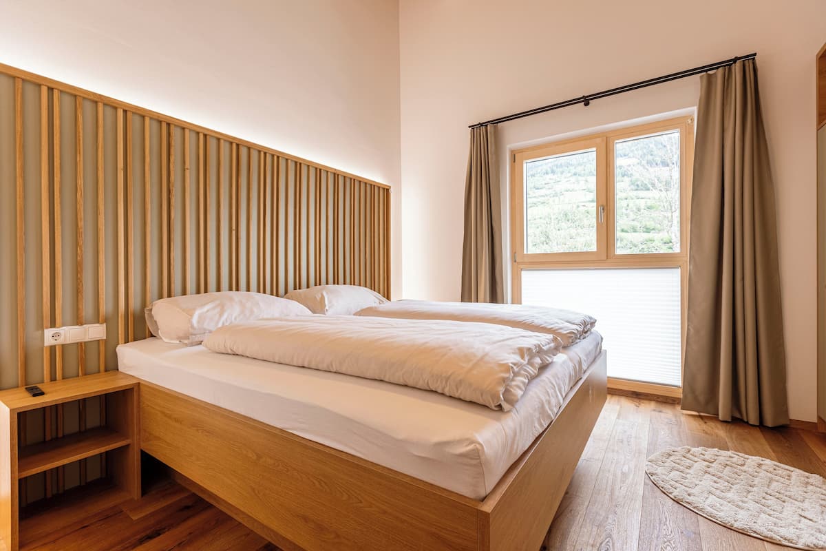 A bedroom with a wooden bed, a nightstand, a window with curtains, and a view of greenery outside.