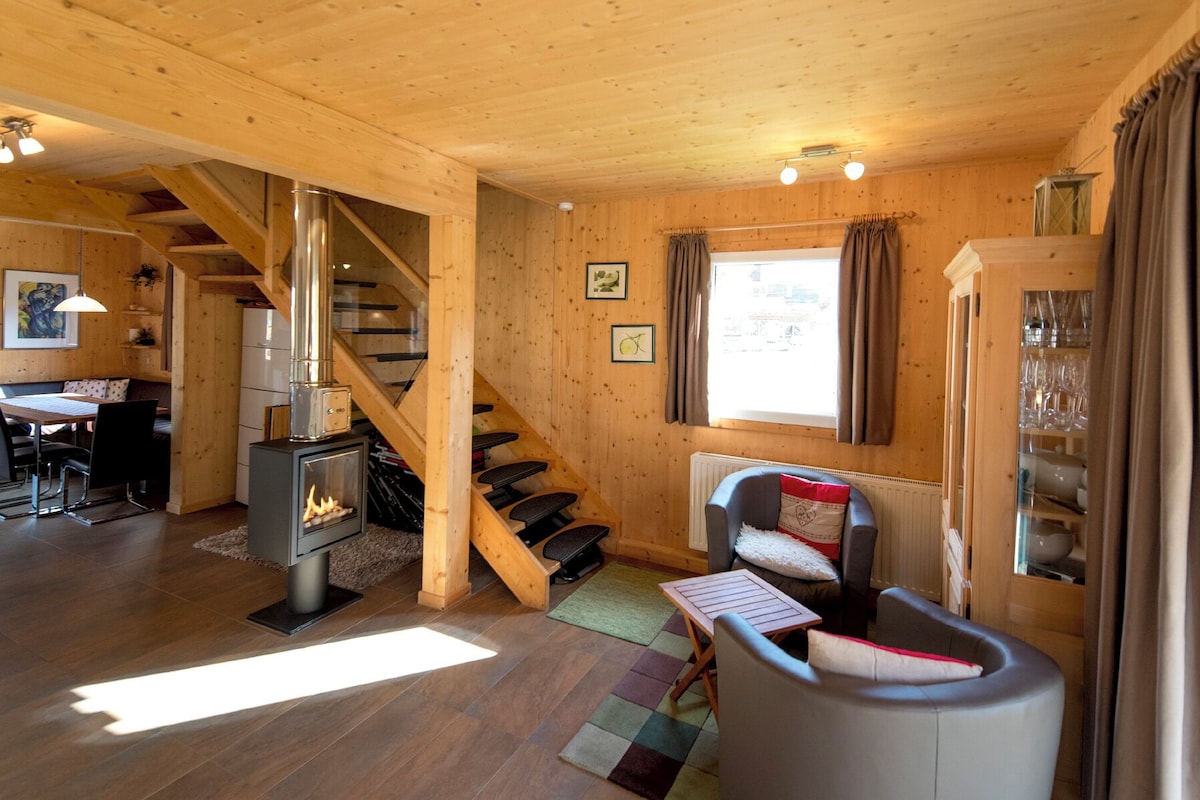 A cozy cabin interior with a wooden staircase, a dining area, a seating area with a table and chairs, and a fireplace.