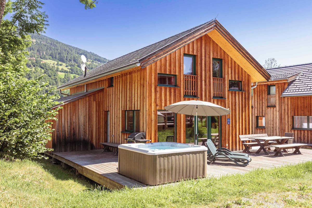 A wooden cabin with a hot tub, outdoor seating, and a deck overlooking a mountainous landscape.