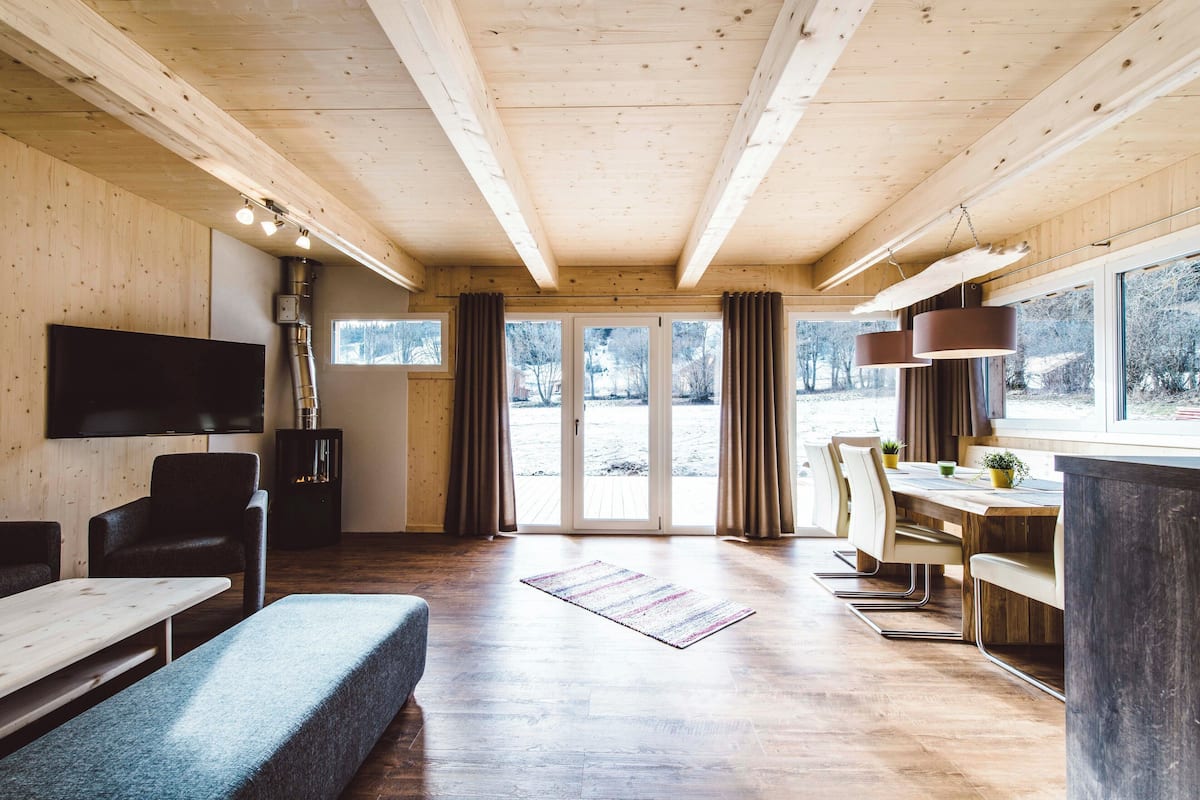 A spacious room with a wooden ceiling, a flat-screen TV, a sofa, a dining table with chairs, and a large window with curtains.