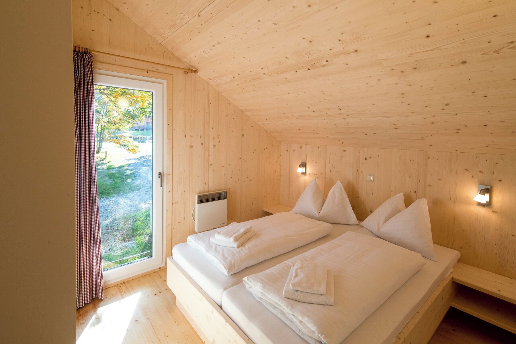 A wooden cabin bedroom with a bed, two bedside tables, a window with curtains, and a wall-mounted heater.
