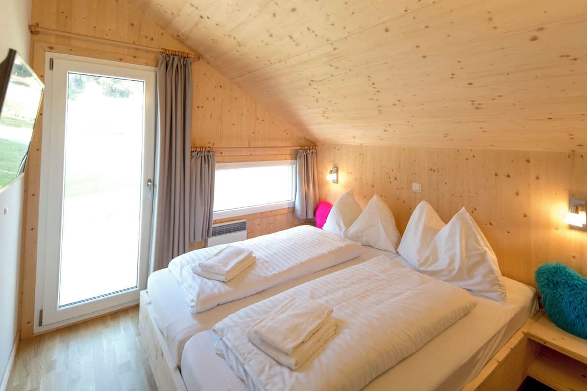 A wooden cabin bedroom with a large bed, two bedside tables, a window with curtains, and a wall-mounted light fixture.
