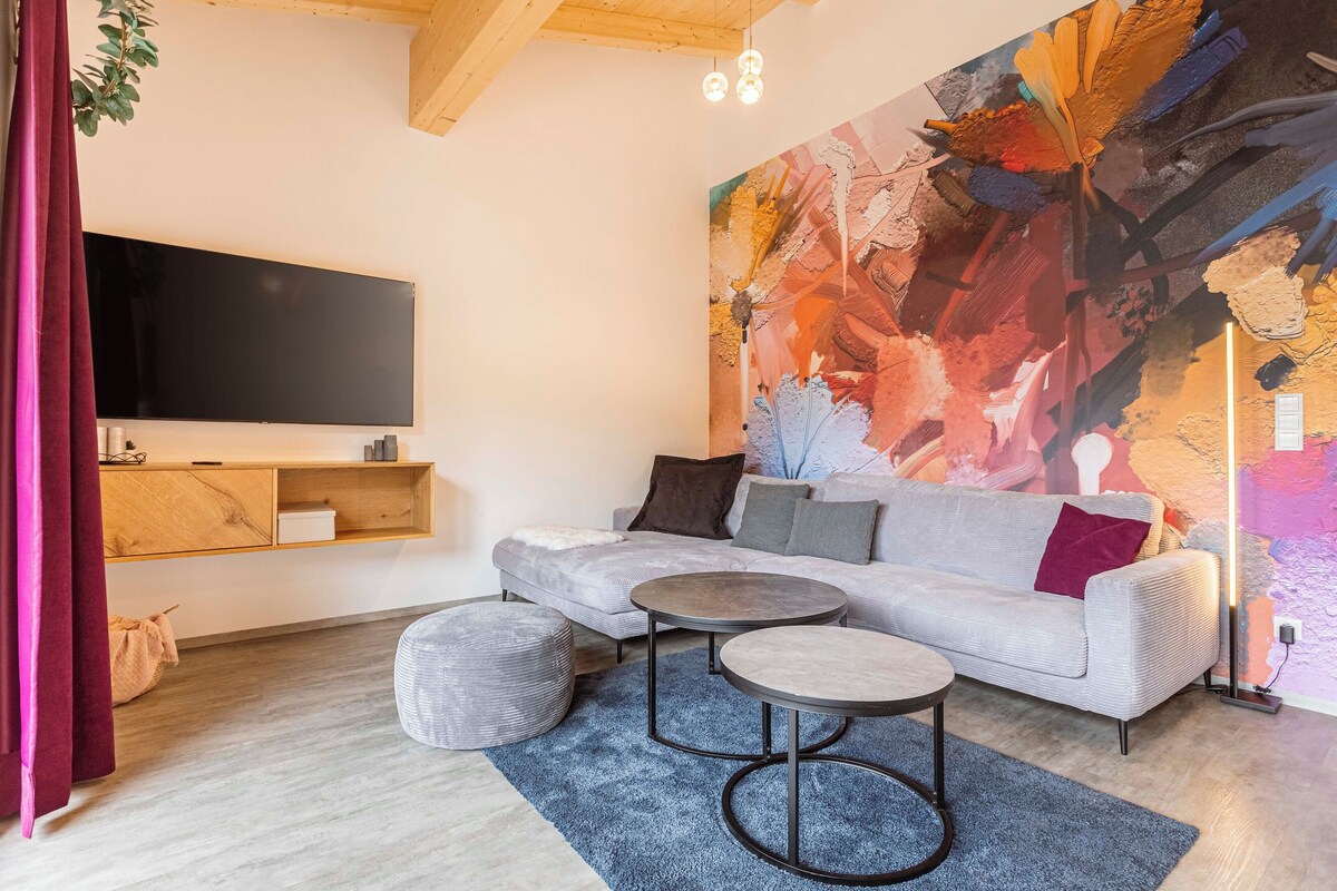 A modern living room with a grey sofa, two round tables, a flat-screen TV, and a vibrant abstract mural.