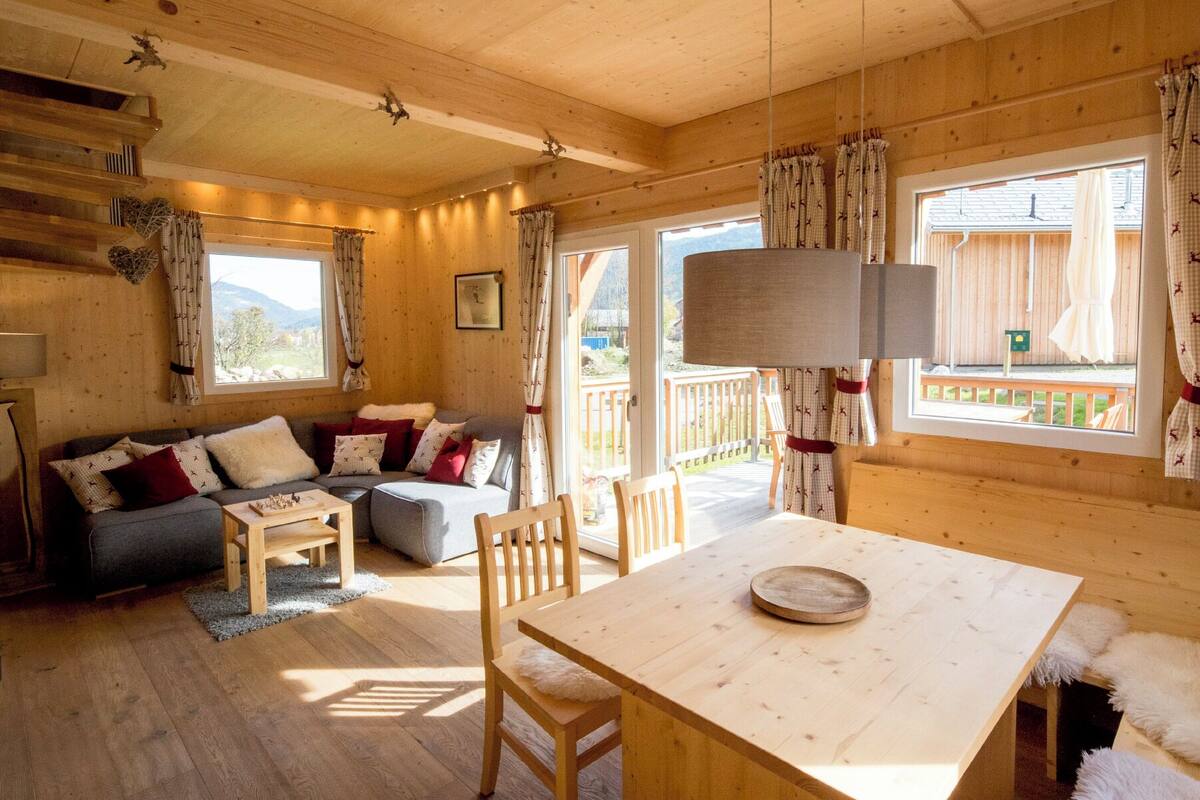 Interior acogedor de una cabaña con una mesa de comedor de madera, un sofá con cojines y vistas a una terraza y montañas a través de ventanas amplias.