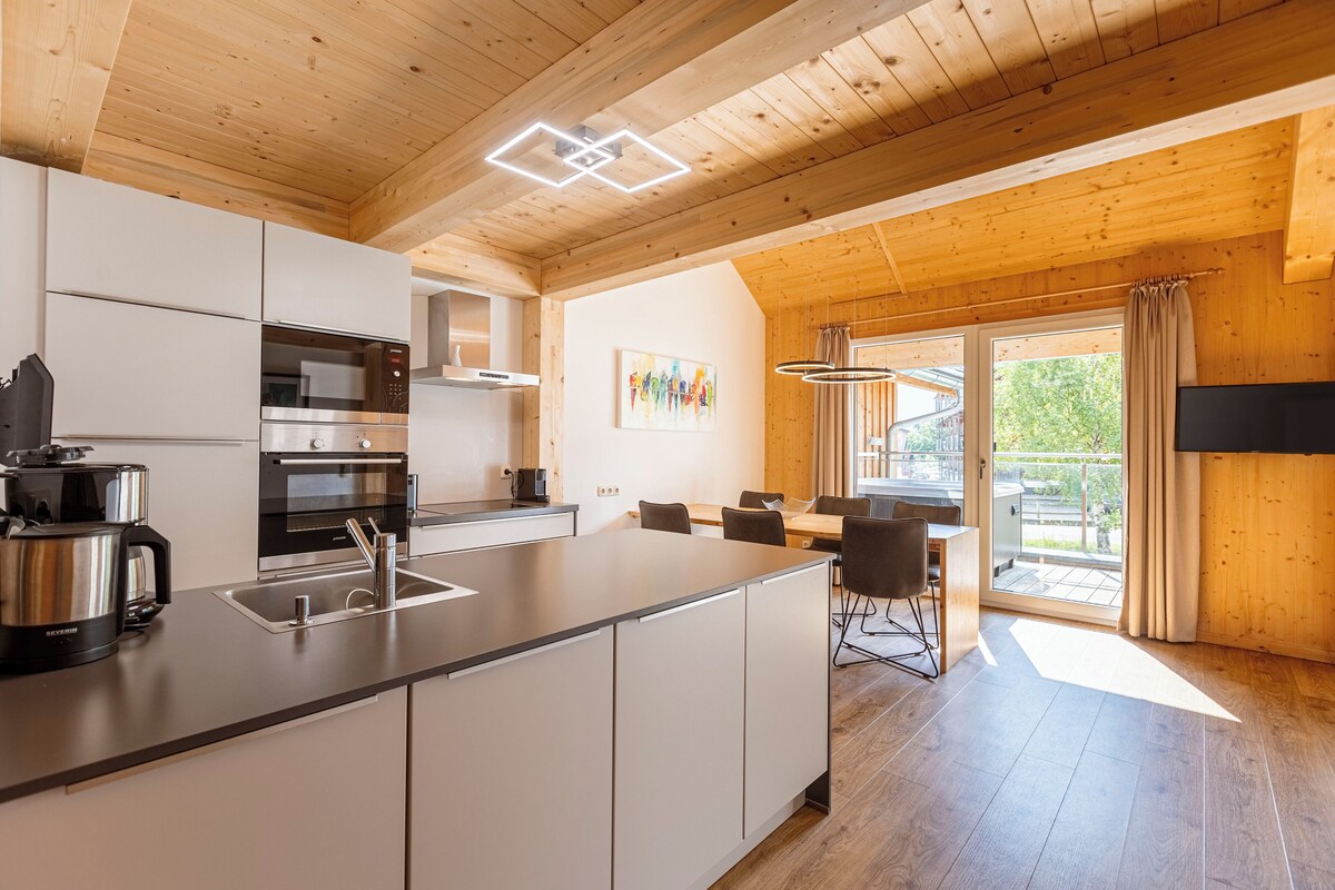 A modern kitchen with white cabinets, stainless steel appliances, and a large island. There is a dining area with a table and chairs, and a large window with curtains.